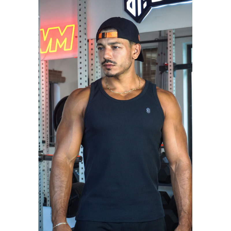 Homme musculé portant débardeur noir ESSENTIAL, strapless cap noir, chaîne dorée; gym intérieure avec équipements en arrière-pla