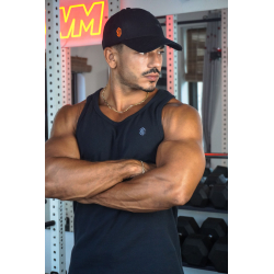 Homme musclé portant un débardeur noir ESSENTIAL, casquette noire, bras croisés, salle de sport avec haltères en arrière-plan.