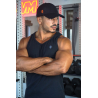 Homme musclé portant un débardeur noir ESSENTIAL, casquette noire, bras croisés, salle de sport avec haltères en arrière-plan.
