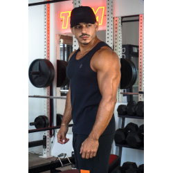 Homme musclé portant débardeur noir ESSENTIAL dans une salle de sport, casquette noire, barres et haltères en arrière-plan, look