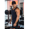 Homme musclé portant débardeur noir ESSENTIAL dans une salle de sport, casquette noire, barres et haltères en arrière-plan, look
