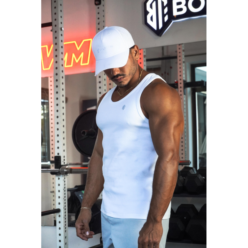Homme musclé portant un débardeur blanc ESSENTIAL et une casquette blanche dans une salle de sport, en train de s’entraîner.