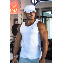 Homme musclé portant débardeur blanc ESSENTIAL dans une salle de sport, casquette blanche et collier doré, décor de gym en arriè