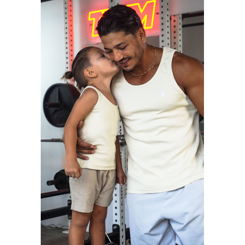 Homme adulte et jeune garçon, tous deux en débardeurs crème, s’embrassant près d’un rack de musculation dans une salle de sport.