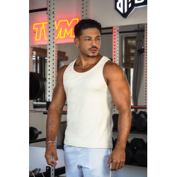 Homme musclé en débardeur crème ESSENTIAL dans une salle de sport, portant collier, près d’haltères et d’un rack de poids.