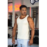 Homme musclé en débardeur crème ESSENTIAL dans une salle de sport, portant collier, près d’haltères et d’un rack de poids.