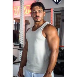 Homme musclé debout dans une salle de sport, porte un débardeur beige, collier doré, en regardant sur le côté.