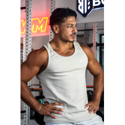 Homme musclé en débardeur ESSENTIAL beige, pose les mains sur les hanches dans une salle de sport, collier doré.