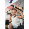 Petit garçon assis sur un équipement de musculation, poings en l’air, debardeur beige ESSENTIAL, intérieur d’une salle de sport.