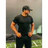 Homme musclé en T-shirt noir ESSENTIAL, casquette noire, pose dans une salle de sport.
