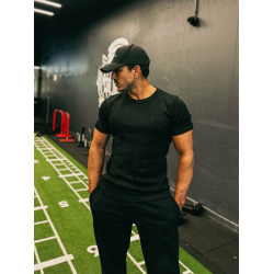 Homme musclé en noir, t-shirt et casquette grimpeur, debout dans salle de sport noire, gazon synthétique et matériel d’entraînem