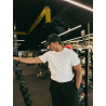 Homme sportif en T-shirt blanc ESSENTIAL, noir pantalon et casquette noire, debout dans une salle de sport près d’un rack d’halt