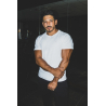 Homme musclé en t-shirt blanc ESSENTIAL, pose les bras croisés dans un décor sombre.