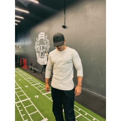 Homme musclé portant T-shirt manches longues beige et casquette noire dans une salle de sport moderne.