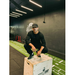 Homme musclé en tenue noire s’agenouillant sur un wooden plyo-box dans une salle de sport, casquette noire, décor gris et logo F