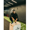 Homme musclé en tenue noire s’agenouillant sur un wooden plyo-box dans une salle de sport, casquette noire, décor gris et logo F