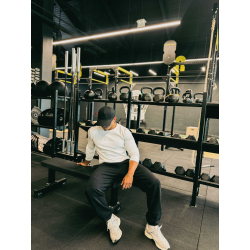 Homme assis sur un banc de salle de sport, portant t-shirt blanc à manches longues et casquette noire, entouré d'haltères et de
