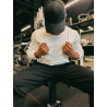 Homme assis sur un banc de musculation, portant un t‑shirt blanc ESSENTIAL manches longues et une casquette noire, dans une sall
