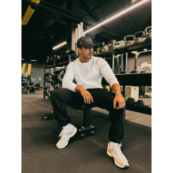 Homme athlétique assis sur un banc de gym, portant un t-shirt blanc à manches longues, casquette noire, pantalon noir et sneaker