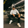 Homme athlétique assis sur un banc de gym, portant un t-shirt blanc à manches longues, casquette noire, pantalon noir et sneaker