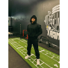 Homme en hoodie noir et casquette blanche, debout sur une surface verte en gazon artificiel dans une salle de sport, bras croisé