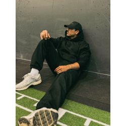 Homme vêtu de noir assis par terre contre un mur gris, bonnet et hoodie, baskets blanches, sur une surface de sport avec gazon a
