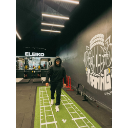 Homme en noir avec capuche marche sur une piste de sprint verte dans une salle de sport, mur décoratif et logos Eleiko en arrièr
