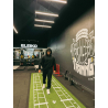 Homme en noir avec capuche marche sur une piste de sprint verte dans une salle de sport, mur décoratif et logos Eleiko en arrièr