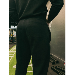 Homme vêtu d'un pantalon noir ajusté vu de dos dans une salle de sport, près d'un mur et d'équipements; arrière-plan partielleme