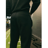 Homme vêtu d'un pantalon noir ajusté vu de dos dans une salle de sport, près d'un mur et d'équipements; arrière-plan partielleme