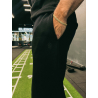 Homme en pantalon noir près d’un entraîneur, main dans la poche, bracelet doré; logo discret sur le pantalon. Jogging ESSENTIAL
