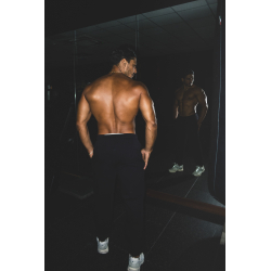 Homme musclé debout torse nu dans une salle, vu de dos, se regardant dans un miroir, portant pantalon noir et baskets blanc; amb