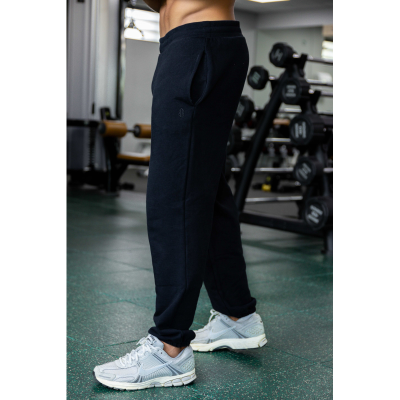 Personne debout dans une salle de sport, pantalon jogging noir ESSENTIAL noir et baskets grises Nike, sur un sol vert, matériel
