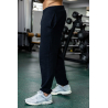 Personne debout dans une salle de sport, pantalon jogging noir ESSENTIAL noir et baskets grises Nike, sur un sol vert, matériel