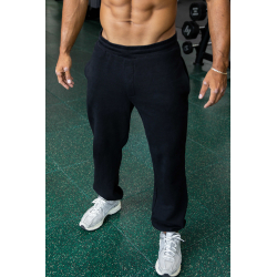 Homme musclé debout en pantacourt noir et baskets gris, en salle de sport avec poids en arrière-plan.