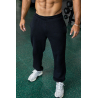 Homme musclé debout en pantacourt noir et baskets gris, en salle de sport avec poids en arrière-plan.