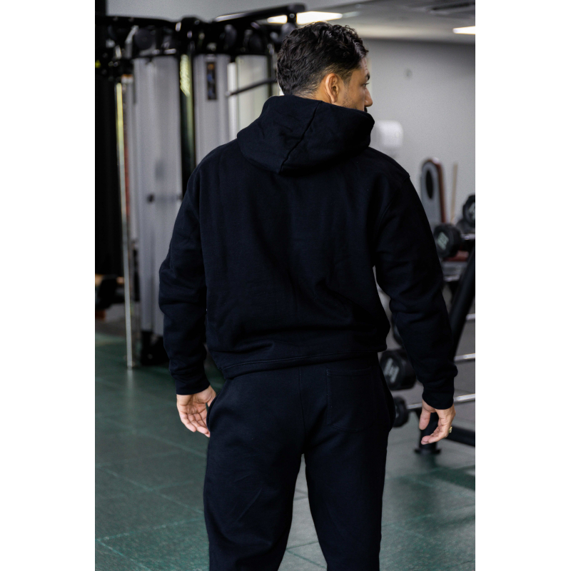 Homme vu de dos portant un hoodie noir et pantalon assorti, dans une salle de sport avec appareils de musculation.