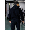 Homme vu de dos portant un hoodie noir et pantalon assorti, dans une salle de sport avec appareils de musculation.