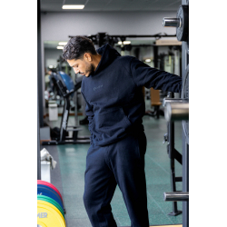 Homme sportif en tenue noir, debout dans une salle de sport, appuyé contre une machine, regard vers le sol.