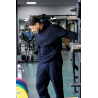 Homme sportif en tenue noir, debout dans une salle de sport, appuyé contre une machine, regard vers le sol.