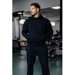 Homme athlétique en noir, hoodie et joggers noir, debout dans une salle de sport avec matériel de musculation en arrière-plan.