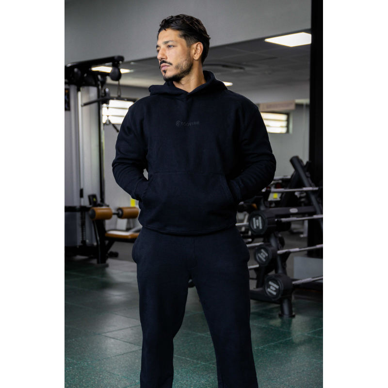 Homme athlétique en noir, hoodie et joggers noir, debout dans une salle de sport avec matériel de musculation en arrière-plan.