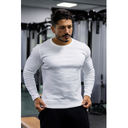 Homme musclé portant T-shirt blanc à manches longues ESSENTIAL dans une salle de sport, fond machines et éclairage flou.