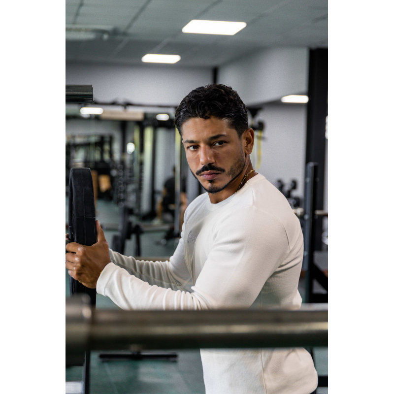 Homme musclé en haut blanc à manches longues, debout près d’un appareil de musculation dans une salle de sport.