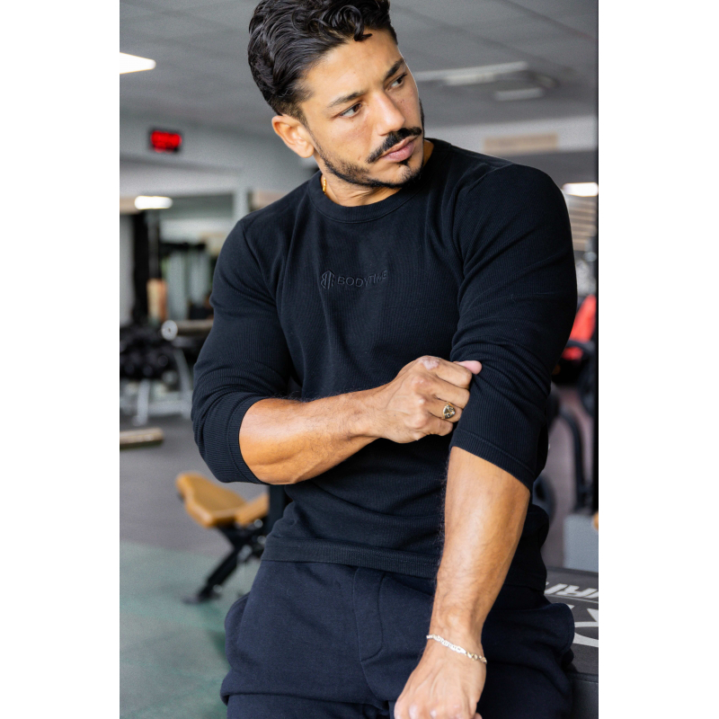 Homme musclé en t-shirt noir à manches longues, regard sur le bras, intérieur de salle de sport. Logo “BODYTIME” sur chest.