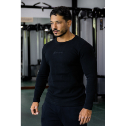 Homme musclé dans une salle de sport, chemise noire à manches longues ESSENTIAL noir, regard posé, chaîne dorée, ambiance gym.