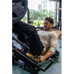 Homme musclé en T-shirt beige à manches longues, assis sur une machine de musculation, engageant les jambes sur une presse, gym