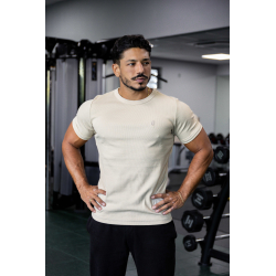 Homme musclé dans une salle de sport portant un t-shirt esssential beige clair, main sur les hanches, arrière-plan de matériel d