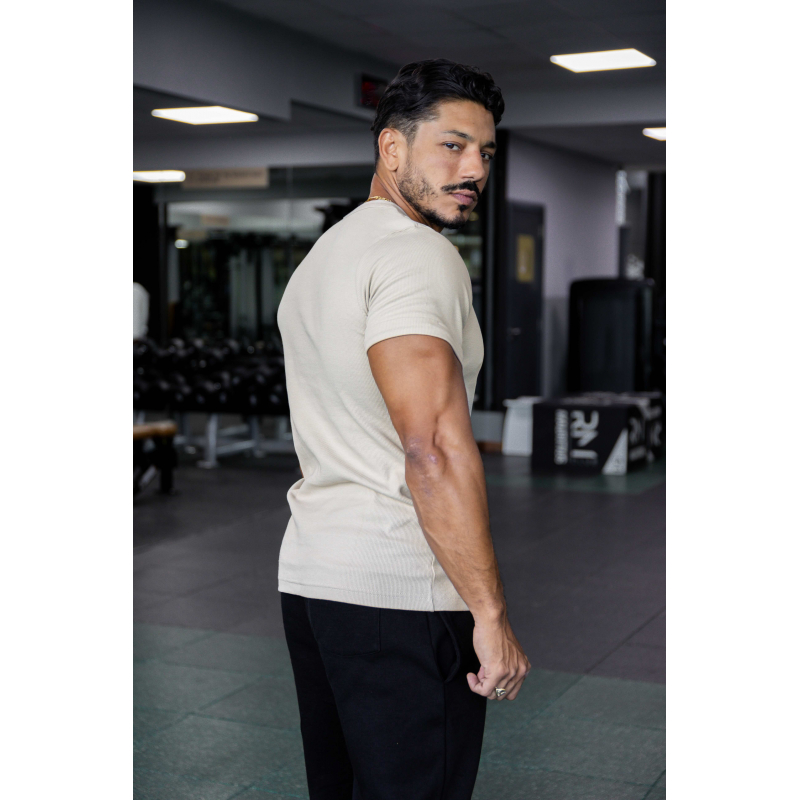 Homme musculé en t-shirt beige ESSENTIAL, vue de profil dans une salle de sport, portant pantalon noir.