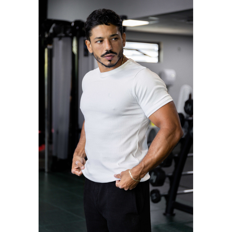 Homme musclé portant un t-shirt blanc ESSENTIAL, près d’un équipement de gym, look soigné et bandeau sur la poitrine.